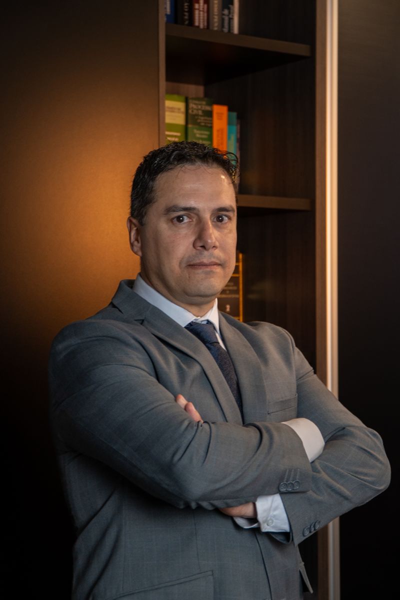 Retrato do sócio Rodrigo Dal Forno de Camargo, de terno cinza claro e braços cruzados, em ambiente com estante de livros.