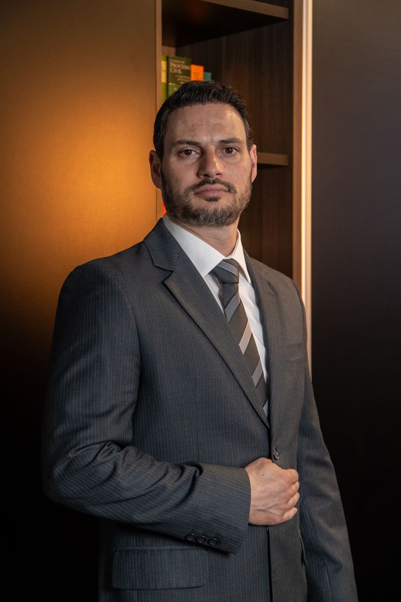 Retrato do sócio Carlos A. Giovaneli Pereira Jr., de barba, terno risca de giz e mão no paletó, em ambiente com estante de livros.